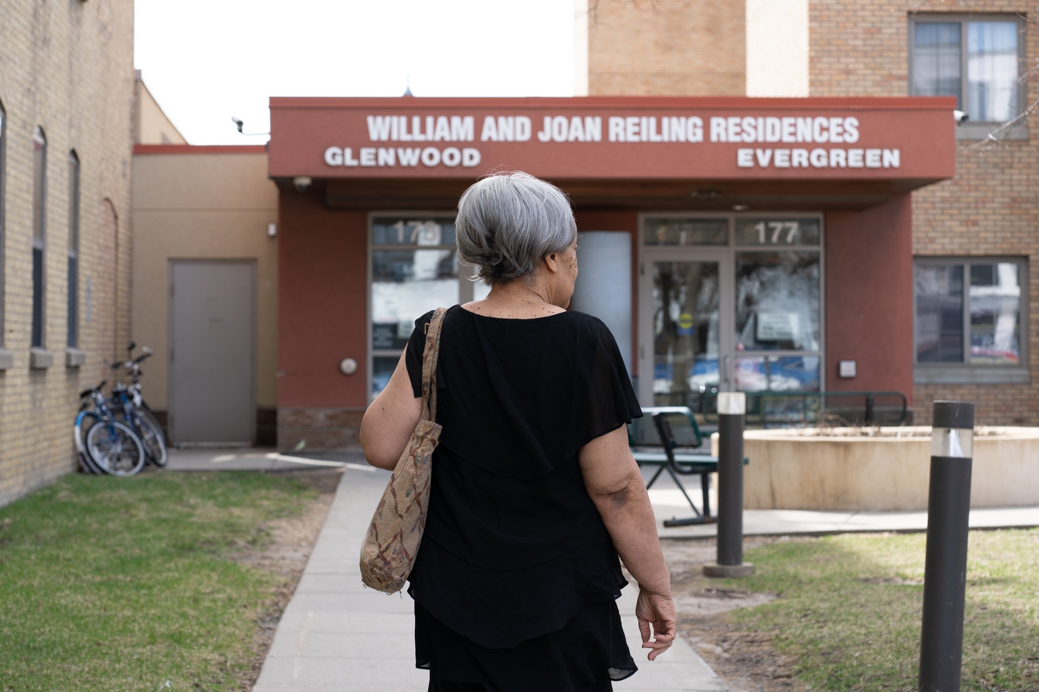 An elderly Black woman with gray hair wearing a black shirt carrying a purse walks outside of a Catholic Charities building