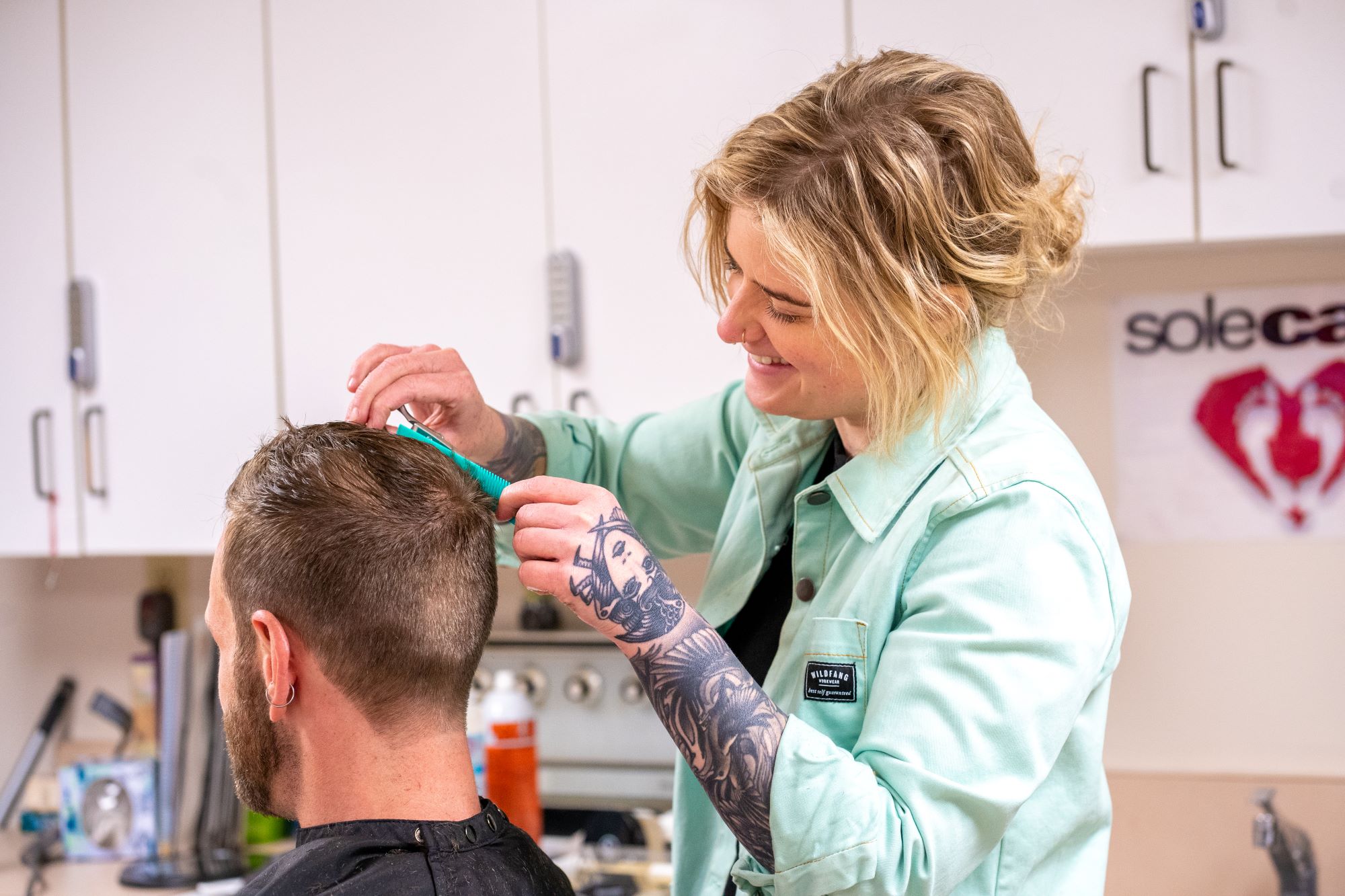 A blonde woman with a tattooed arm cuts a man's hair