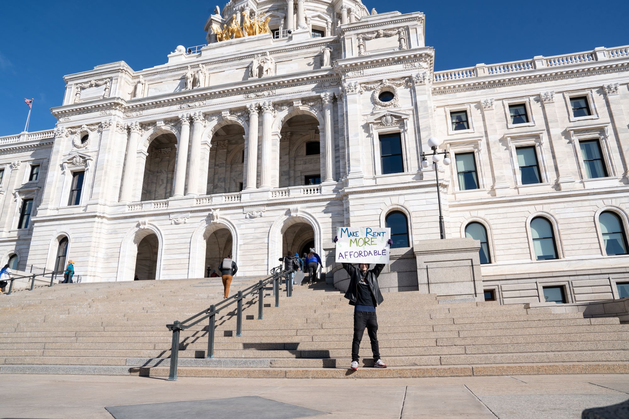 A young man stands in front of the Minnesota Capitol building holding a sign that says "Affordable housing"