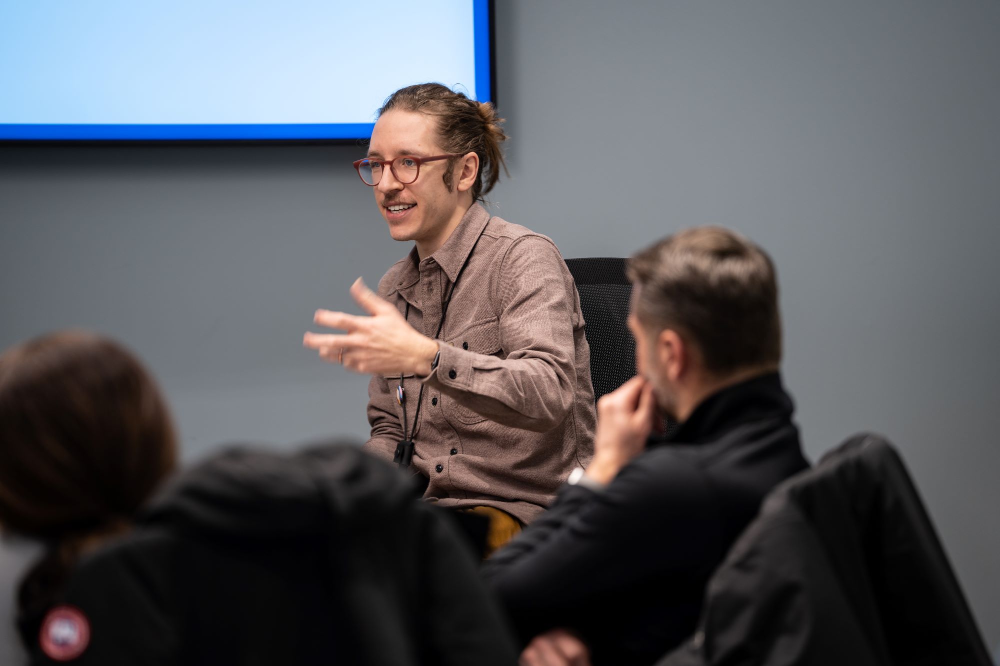 A young man with glasses speaks to a room of people in front of a projector screen