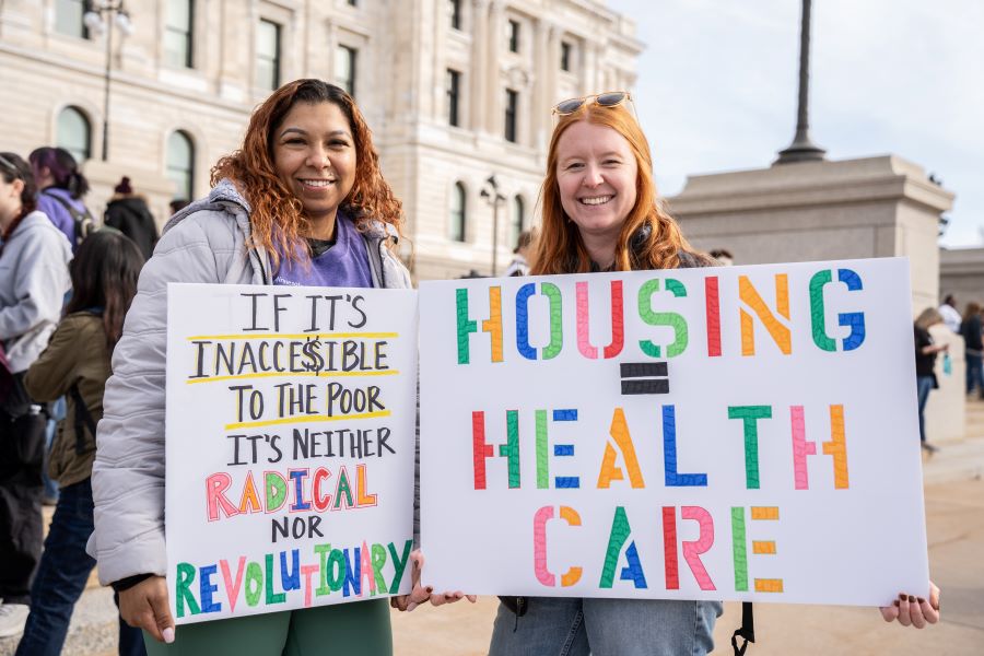 Two red-haired women with signs promoting accessibility and resources for the underprivileged